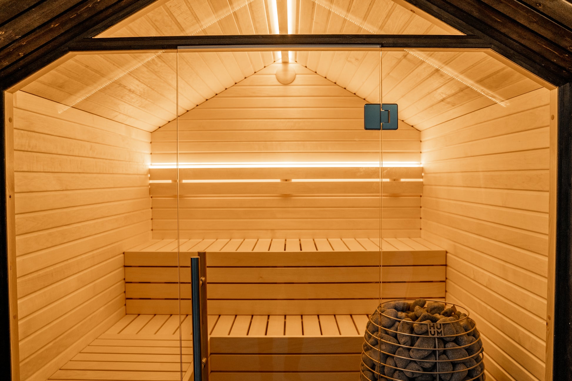 Sauna LUMI con pared de cristal y cesta de piedra.