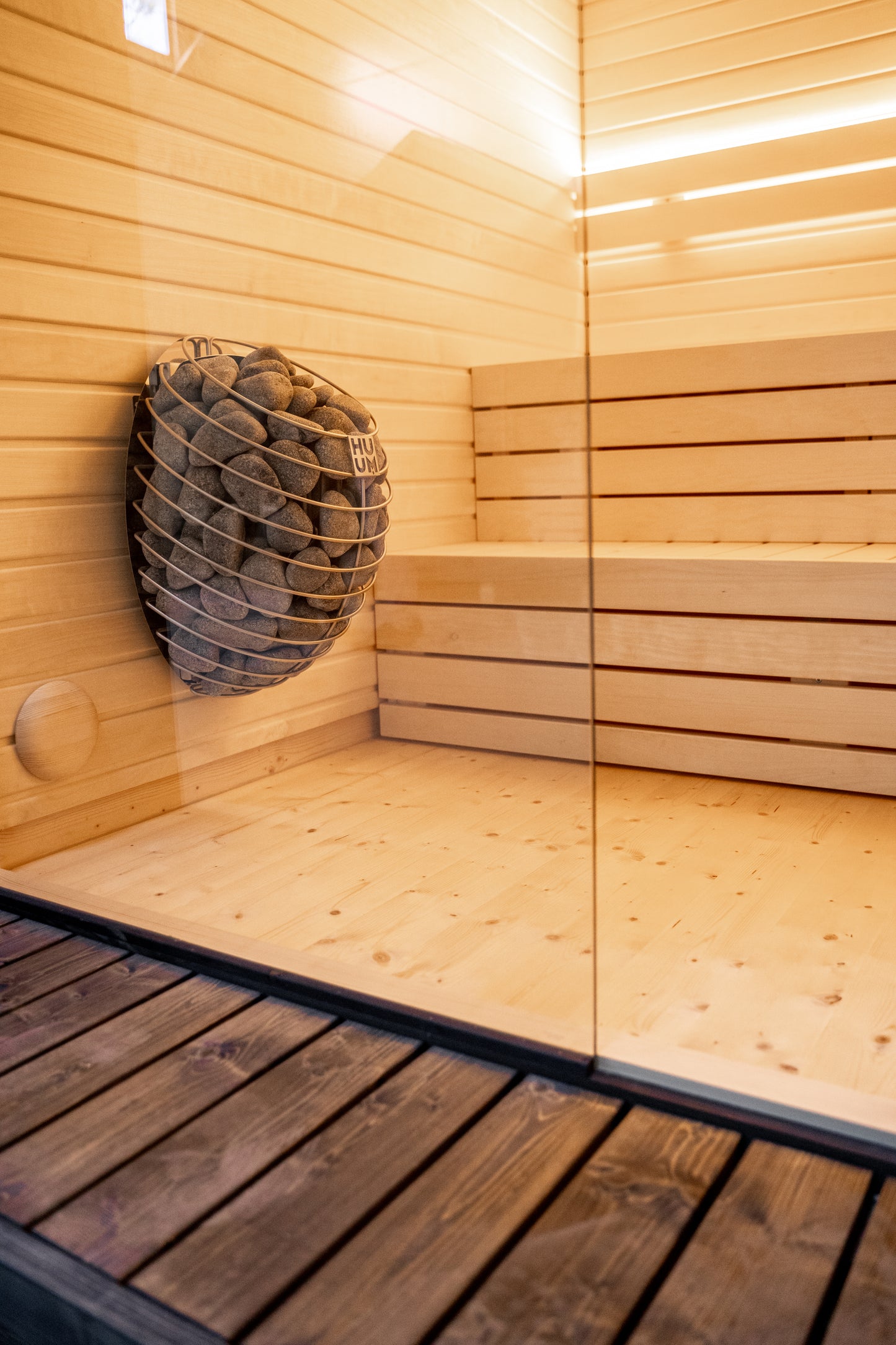 Sauna LUMI con cesta de piedra y pared de cristal.