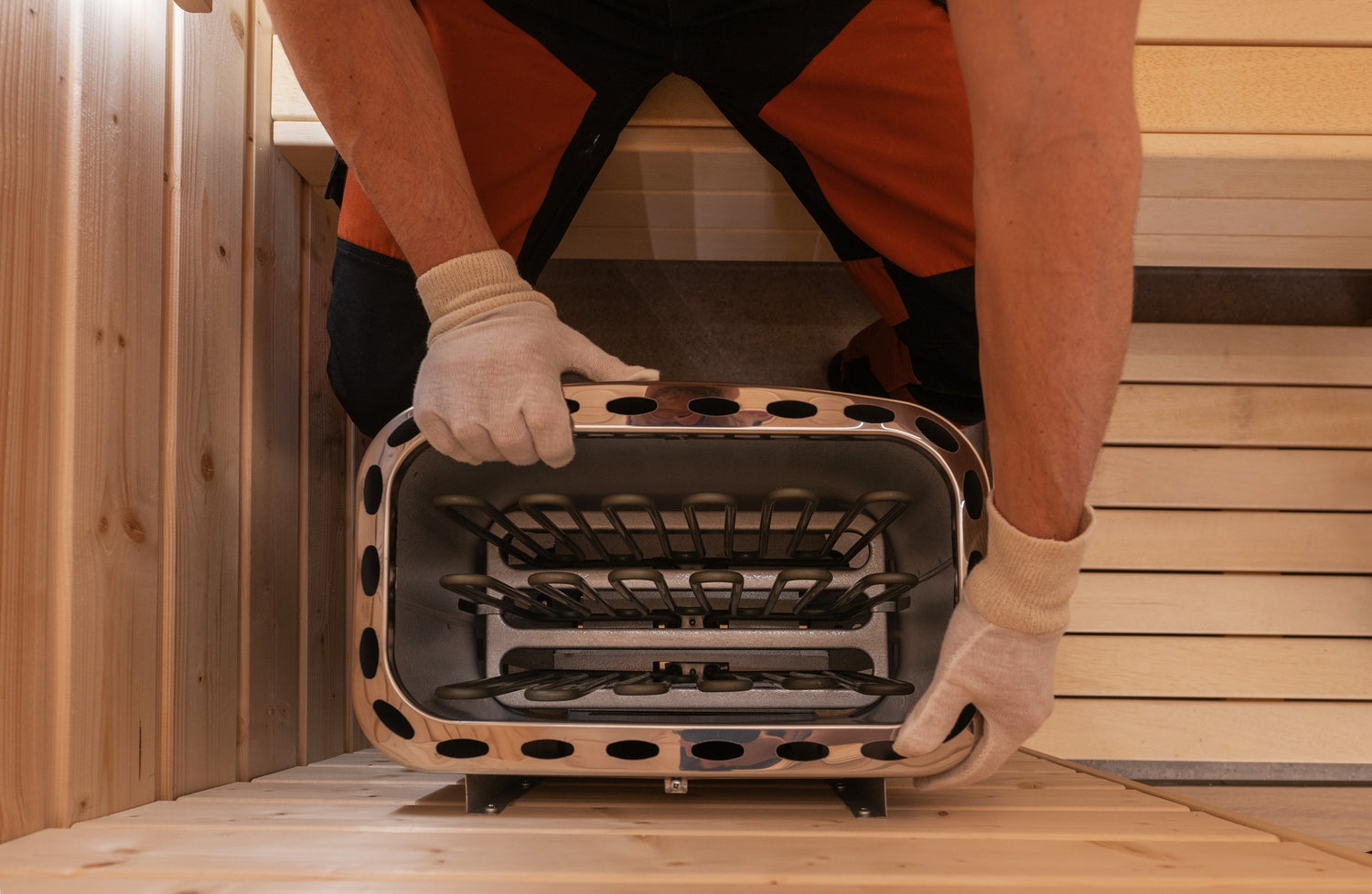 Una persona con pantalones de trabajo naranjas y negros y guantes blancos está instalando un calefactor de acero inoxidable en una habitación de madera. La persona está colocando el calentador dentro de una abertura rectangular entre paneles de madera. La habitación parece formar parte de una sauna.