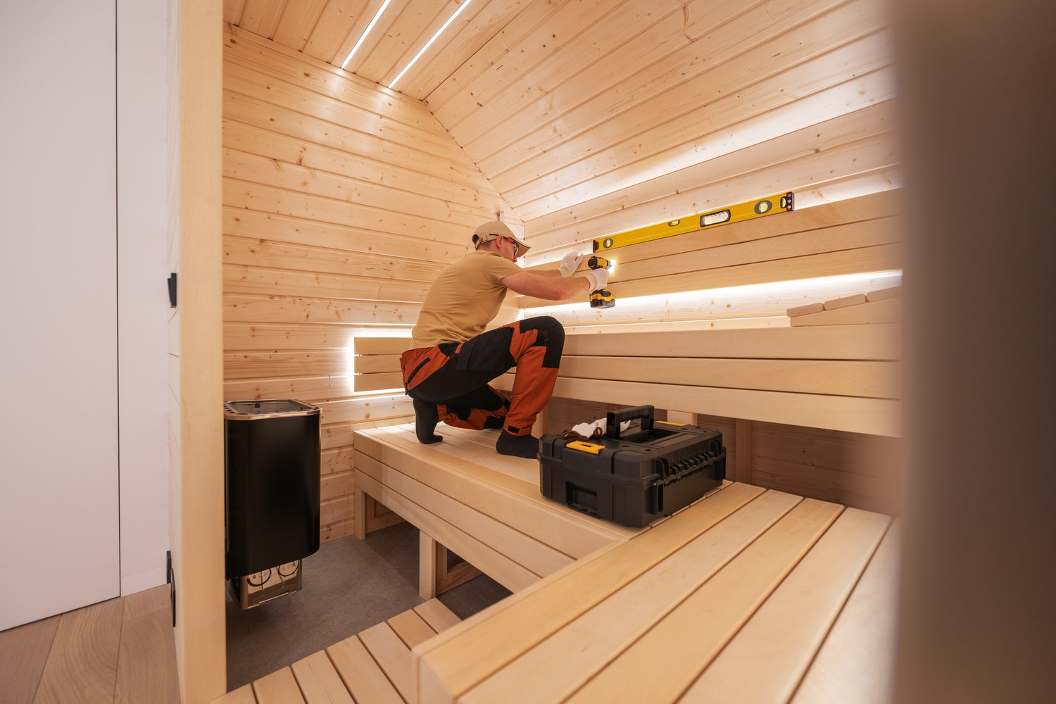 Una persona con camisa beige y pantalones naranjas está trabajando en el interior de una sauna de madera. Está utilizando una herramienta de nivelación para taladrar el respaldo. La sauna tiene bancos y paredes de madera, con una caja de herramientas en uno de los bancos. En una esquina hay un calefactor.